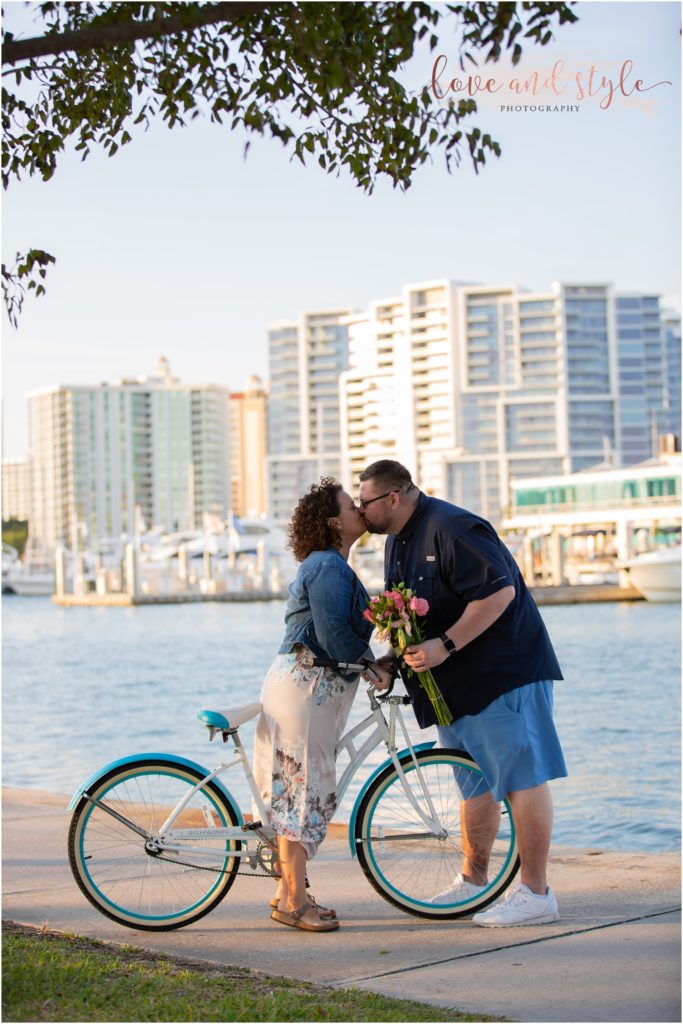 Engagement Photographer Sarasota with couple kissing on a bike with the city behind them
