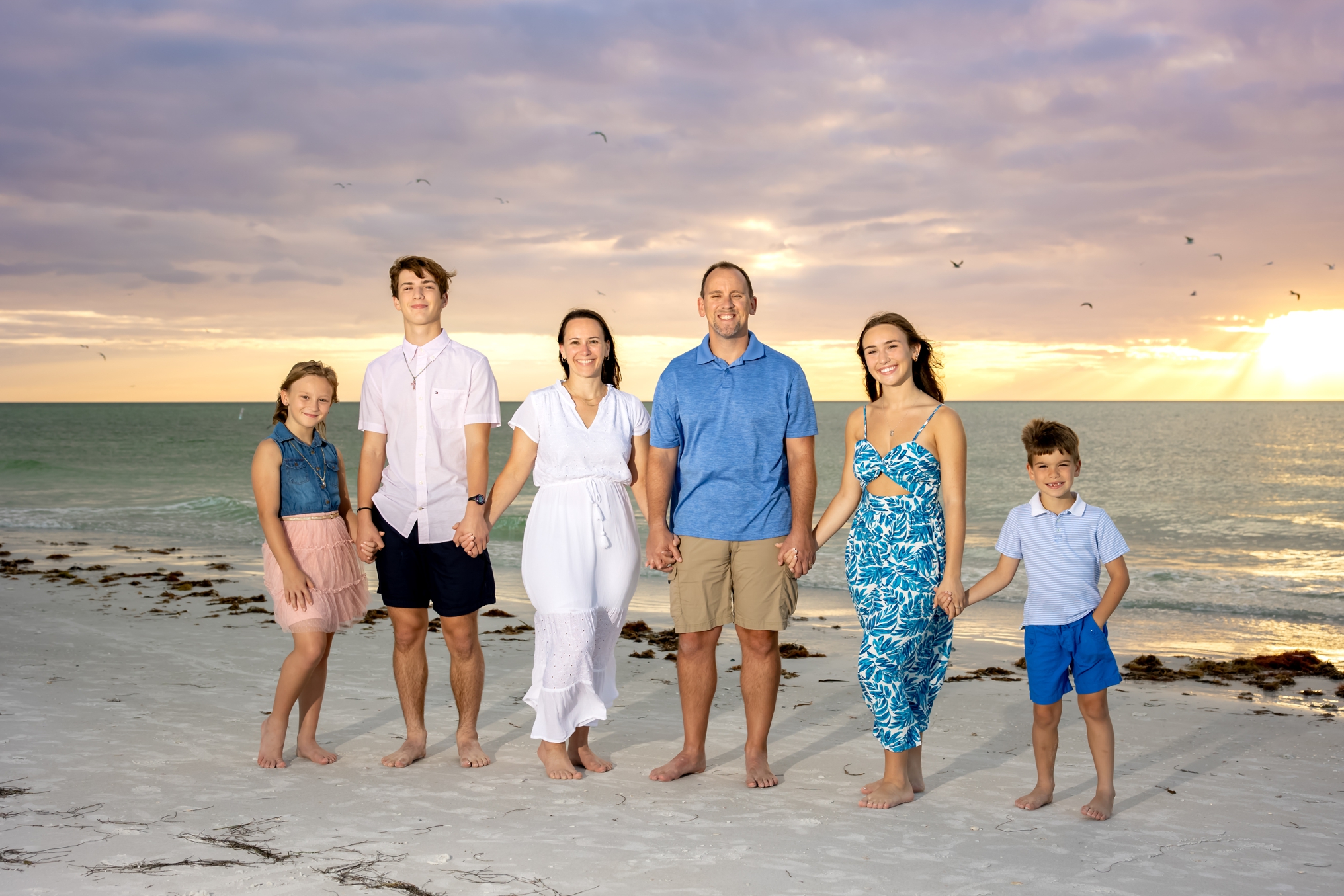 A family of six on the beach holding hands with the sun setting behind them.